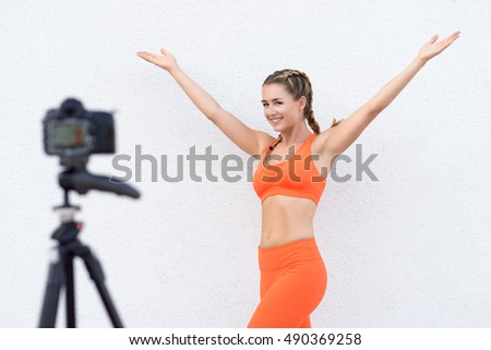 Attractive young video blogger doing a recording filming a fitness exercise and posing with outspread arms and a lovely smile for a camera on a tripod