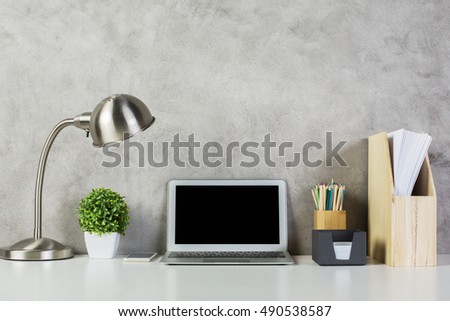 Blank computer on desktop with table lamp, decoratve plant, wooden folder with documents and supplies on concrete wall background. Mock up