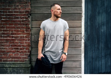 A handsome muscular man wearing blank gray t-shirt is looking aside while standing beside the brick and wooden wall on a street. Empty place for the logo or design. Mock up.