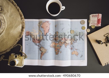 Planning the trip. World map with the hat, film camera, some money, notebook from recycled paper and freshly brewed coffee cup on the dark wooden table background.