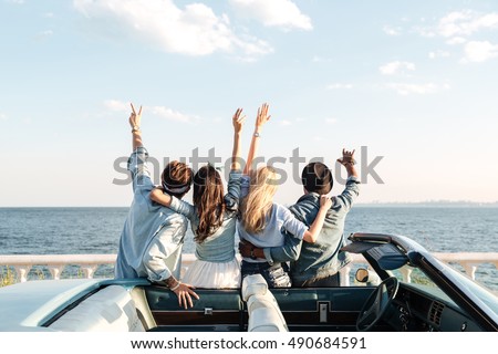 Back view of happy young friends standing with raised hands near the car Royalty-Free Stock Photo #490684591