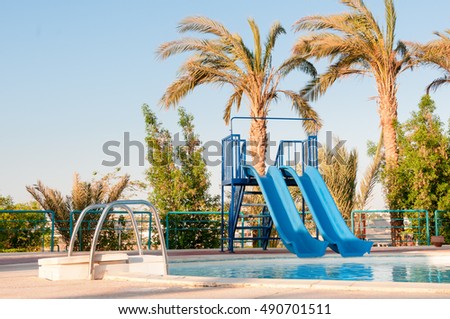 Palm trees sway with two water slides mounted on the pool deck at a luxury, tropical beach resort playground on a sunny day.