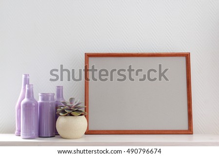 Horizontal blank brown wooden poster frame on white shelf as a part of home decor composition with lilacs glass bottles and succulent plant in a round planter. White texture background.
