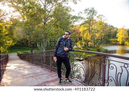 Portrait of the young bearded handsome fashion traveler in the park with casual style and fashion hairstyle and backpack and smart phone on bridge in the beautiful park, posing and enjoy. 
