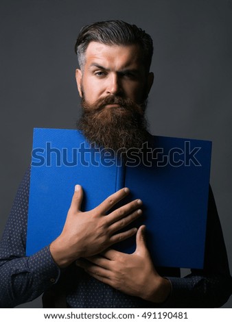 Handsome man hipster with long beard and moustache in shirt holds blue journal on grey background