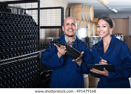 Two friendly smiling  winery workers holding bottle of sparkling wine in aging section of factory
