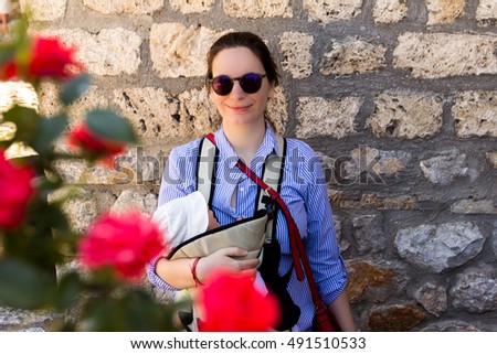 Smiling mother with sunglasses carrying and holding her little baby boy who is sleeping in the carrier with red roses in the front