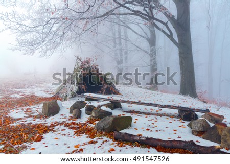 A hut made from leaves and branches stands in a snow covered meadow with trees next to the bench on a misty winter day.