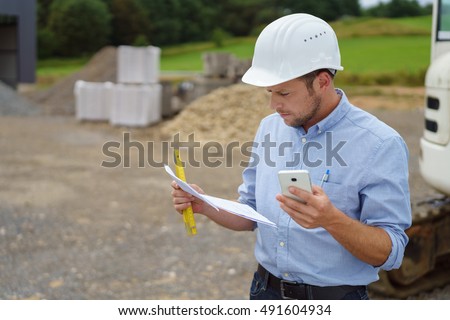Architect or building contractor working on site standing reading a document in his hard hat with a mobile in his hand , with copy space Royalty-Free Stock Photo #491604934