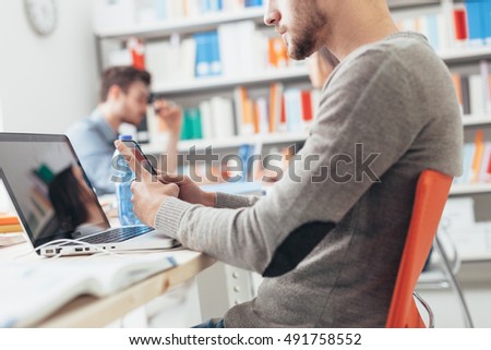 Young college student sitting at desk, using a touch screen smart phone and a laptop, education and technology concept