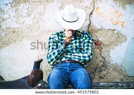 A tough cowboy guy sitting on a wooden fence. He holds his hat, wears jeans and shirt. A brown saddle is next to him. A typical american countryside photo. Royalty-Free Stock Photo #491979802