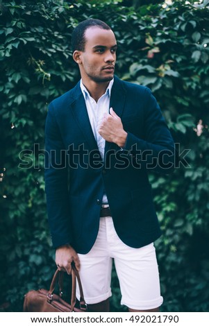 Confident in his style. Handsome young African man in smart casual wear carrying bag and looking away while standing against green plant background outdoors 