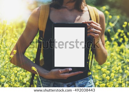Hipster holding in hands tablet computer, mockup of blank empty screen. Girl traveler using gadget on sun flare and yellow flowers background close. Female photographer take view on cell camera
