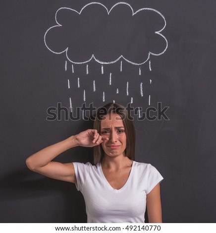 Beautiful young girl is looking at camera and crying, standing against blackboard with drawn rain cloud overhead