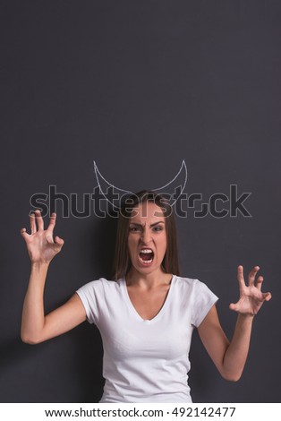 Beautiful young girl is showing devil, showing claws, looking at camera and growling, standing against blackboard with devil's horns overhead