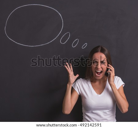 Beautiful young girl is screaming while talking on the mobile phone and gesturing, standing against blackboard with drawn speech bubble