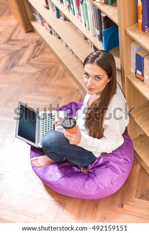 Top view of beautiful student lady studying on laptop computer, drinking coffee and preparing for disciplines or exams in library.