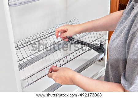 Assembling wall mounted shelf under kitchen cupboard with inside plate rack with drip tray, close-up of a woman hands holds wire dish rack. Royalty-Free Stock Photo #492173068