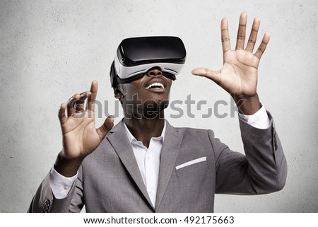 3d technology and virtual reality. Young African dark-skinned businessman dressed in suit playing video game in office using oculus rift headset, smiling and gesturing as if interacting with something