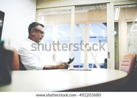 Mature creative news paper editor working on laptop computer at workplace in office meeting room.Male skilled professional writing ideas in notebook after work with start up project on net-book device