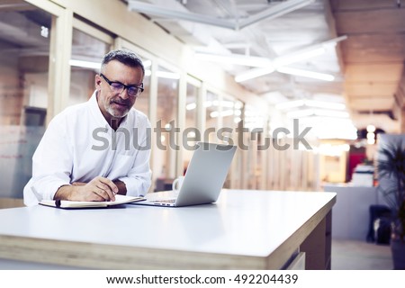 Male skilled jurist writing notes after consultancy with client. Mature editor creating new magazine article by using notebook and laptop computer. Experienced manager developing work-sharing plan Royalty-Free Stock Photo #492204439