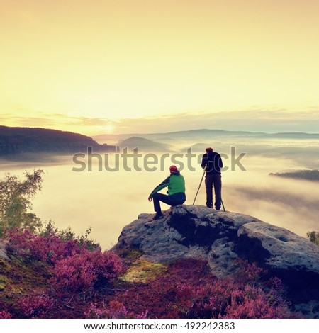 Hiker and photo enthusiast stay with tripod on cliff and takes photos. Dreamy fogy landscape, blue misty sunrise in a beautiful valley below