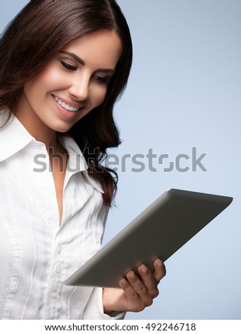 Portrait of happy smiling beautiful young businesswoman using no-name tablet pc, over grey background. Caucasian brunette model in business concept shoot.