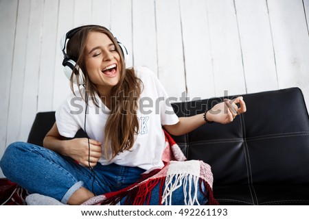 Girl in plaid listening music in headphones, sitting on sofa. Royalty-Free Stock Photo #492261193