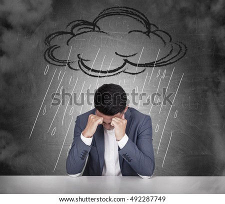Stressed out Asian businessman sitting under cartoon rain cloud in his office. Concept of bad mood.