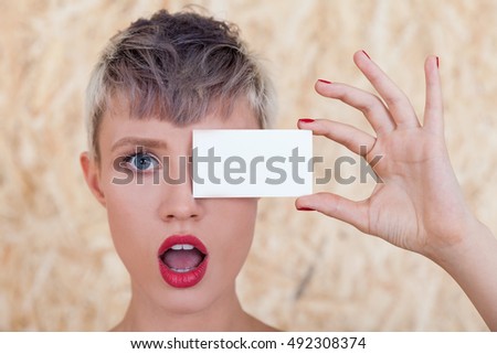 Astonished girl with short hair is closing her eye with blank business card. Concept of insert your ads here. Mock up.