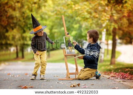 Two sweet children, boy brothers, having fun painting in autumn park together. One child paint the other kid, dressed for halloween