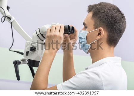 Male dentist looks into the dental microscope which he holds with his hands in the clinic on the wall background. He wears a white uniform and a blue medical mask. Horizontal.