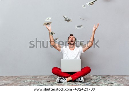 Casual young man sitting on the floor with rain of money over gray background
