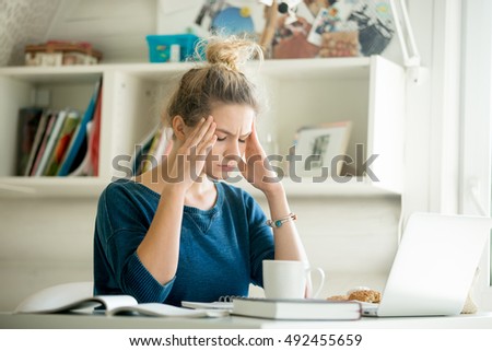 Portrait of an attractive woman at table with cup and laptop, book, notebook on it, hands at her temples. Bookshelf at the background. Concept photo