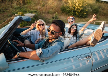 Group of smiling young friends driving car and showing peace sign in summer