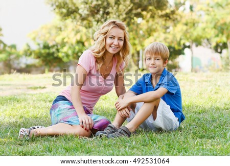Attractive blond woman sitting in the green park with little son.Happy family.Beautiful young adult blonde mother.Smiling white European models.Cute little boy.Togetherness,love and unity concept