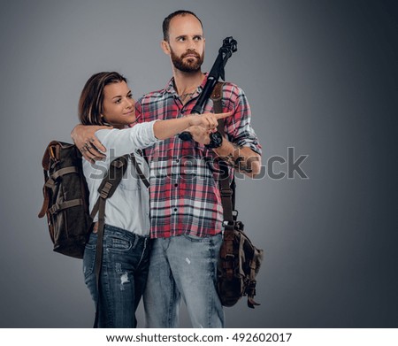 Casual bearded male with photo tripod and backpack posing with attractive urban female on grey background.