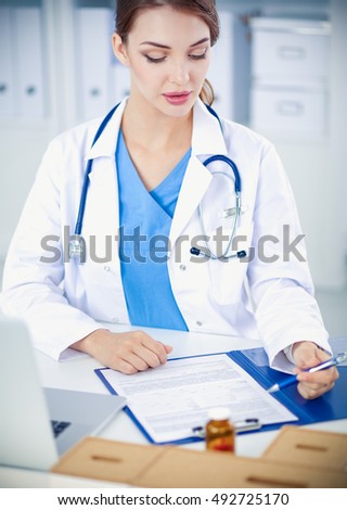 Beautiful young smiling female doctor sitting at the desk and writing.