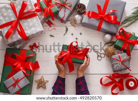 Winter holidays celebrating. Gift wrapping. Packaging modern christmas present boxes in paper with satin red ribbon. Top view of hands on white wood table with fir tree branches, decoration of gift.