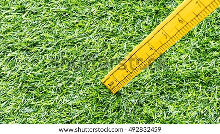Yellow wooden ruler with centimetre and inch indicator on green synthetic grass. Concept of land survey or mapping. Slightly de-focused and close-up shot. Copy space.