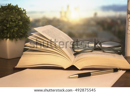 Closeup of open book, glasses, paper sheet, pen and decorative plant on blurry background with city view and sunlight