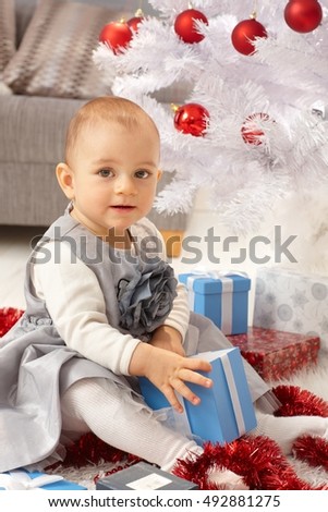 Cute little girl sitting by christmas tree, opening present, smiling.