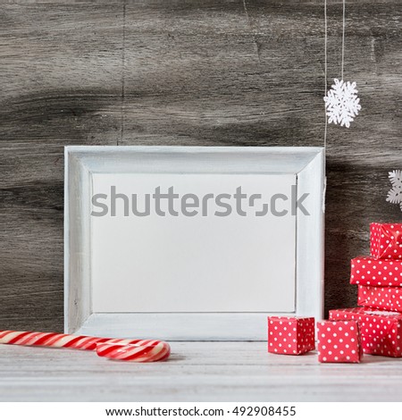 White photo frame with christmas decoration on the wooden table. Red boxes and snowflakes on the grey background. Frame for quotes. Christmas postcard.