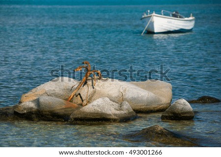 Wooden fishing boat floating on the colourful water. Cyclades, Greece. Beautiful seascape, large old anchor on a stone in the sea, boat in the background.
