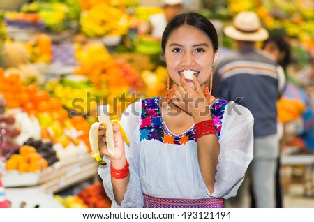 Beautiful young hispanic woman wearing andean traditional blouse posing for camera while eating banana inside fruit market, colorful healthy food selection in background