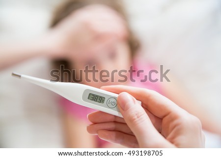 Close-up thermometer.  Mother measuring temperature of her ill kid. Sick child with high fever laying in bed and mother holding thermometer. Hand on forehead.  Royalty-Free Stock Photo #493198705