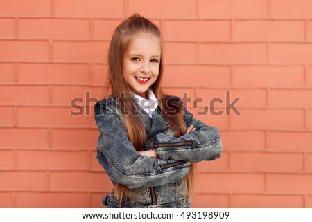 Pretty little girl keeping arms crossed and smiling while standing in front of the brick wall