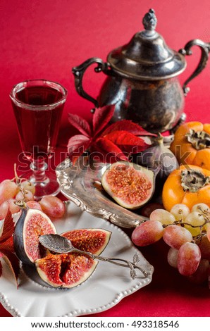 Cut ripe figs on a white vintage plate. The brandy in the glass. Persimmon, figs, grapes on a silver platter. Silverware. Vintage. Autumn crimson leaves. The red background.