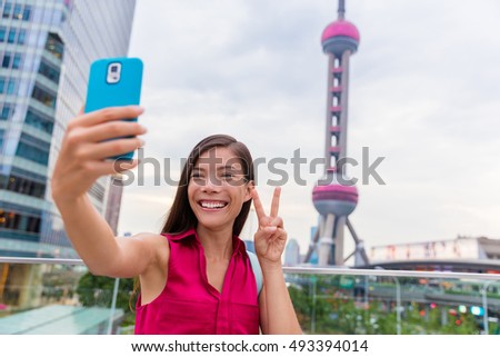 Asian tourist girl taking a selfie smartphone picture at a famous Shanghai landmark in the financial district of Pudong in China. Chinese woman doing a stereotypical japanese v sign for phone photo.