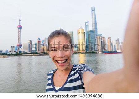 Happy Asian tourist woman taking selfie on The Bund. Smiling young lady holding smartphone camera to take a picture of herself in front of Shanghai's landmak, skyline of skycrapers in Pudong, China.